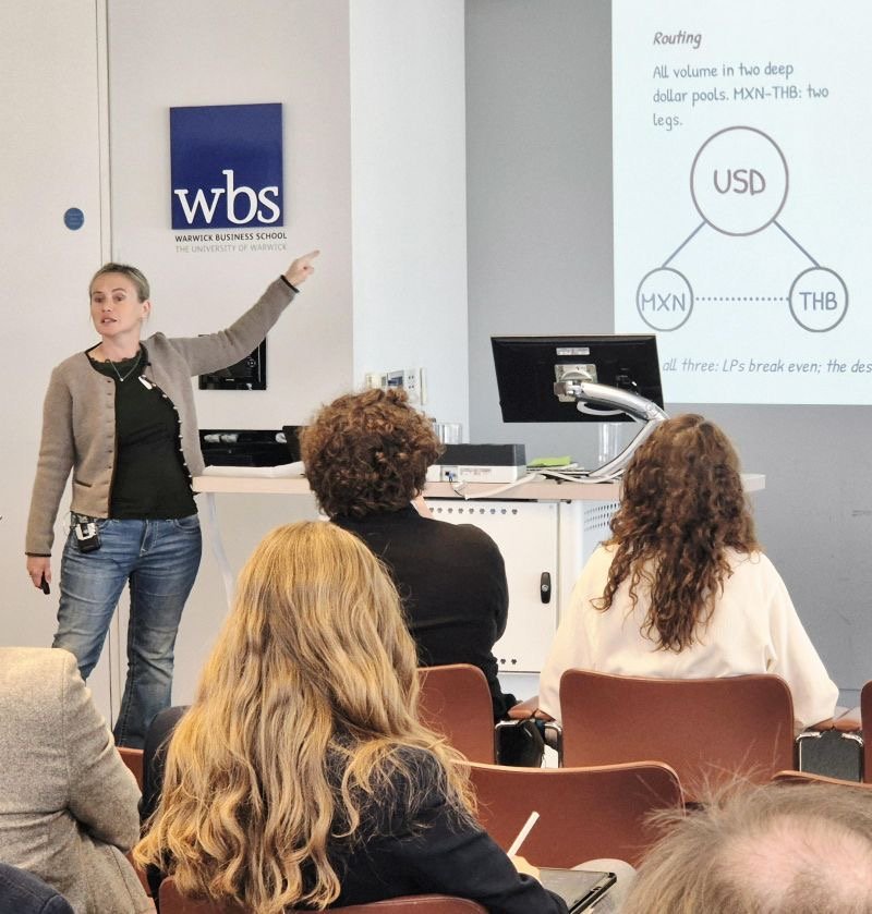 Katya Malinova presenting &ldquo;Dethroning the Dollar?&rdquo; at Warwick Business School, pointing to a slide on routing trades through the U.S. dollar